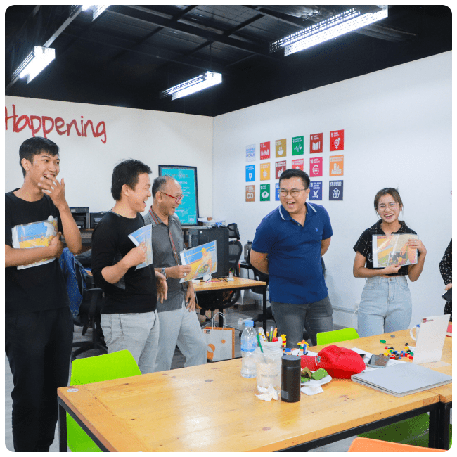 Six people stand in a row around a table laughing during a bootcamp for the SAAMBAT program.