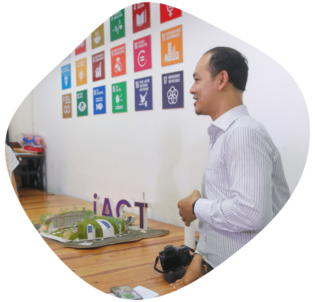 A male entrepreneur stands in front of a mini model of the iACT school, with SDG cards decorating the wall behind him at Impact Hub Phnom Penh.