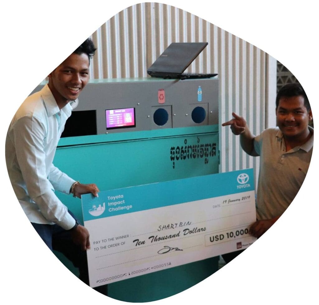 Two entrepreneurs, founders of SmartBin, stand with an oversized check in front of their prototype commercial trash and recycling machine.