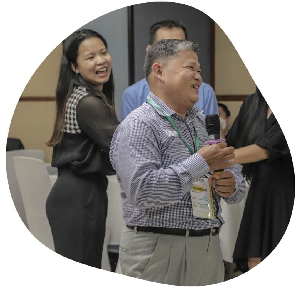 A Cambodian university professor stands with a microphone laughing. A few people are visible behind him also laughing.