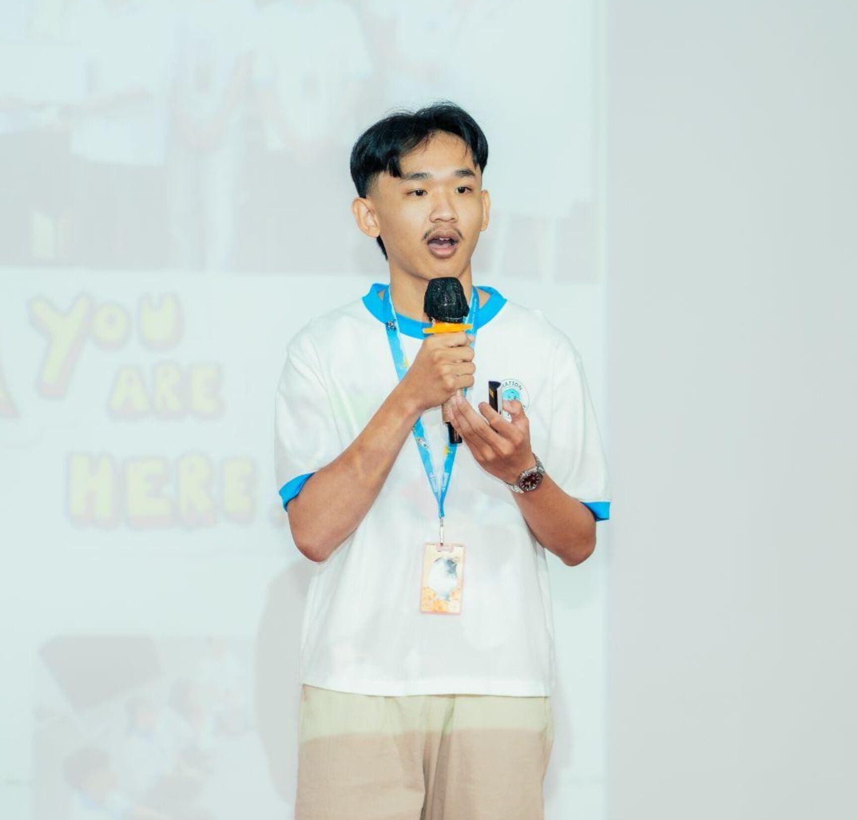 Young man speaking into a microphone on stage, wearing a lanyard and white shirt. Background shows blurred text. Confident and engaged tone.