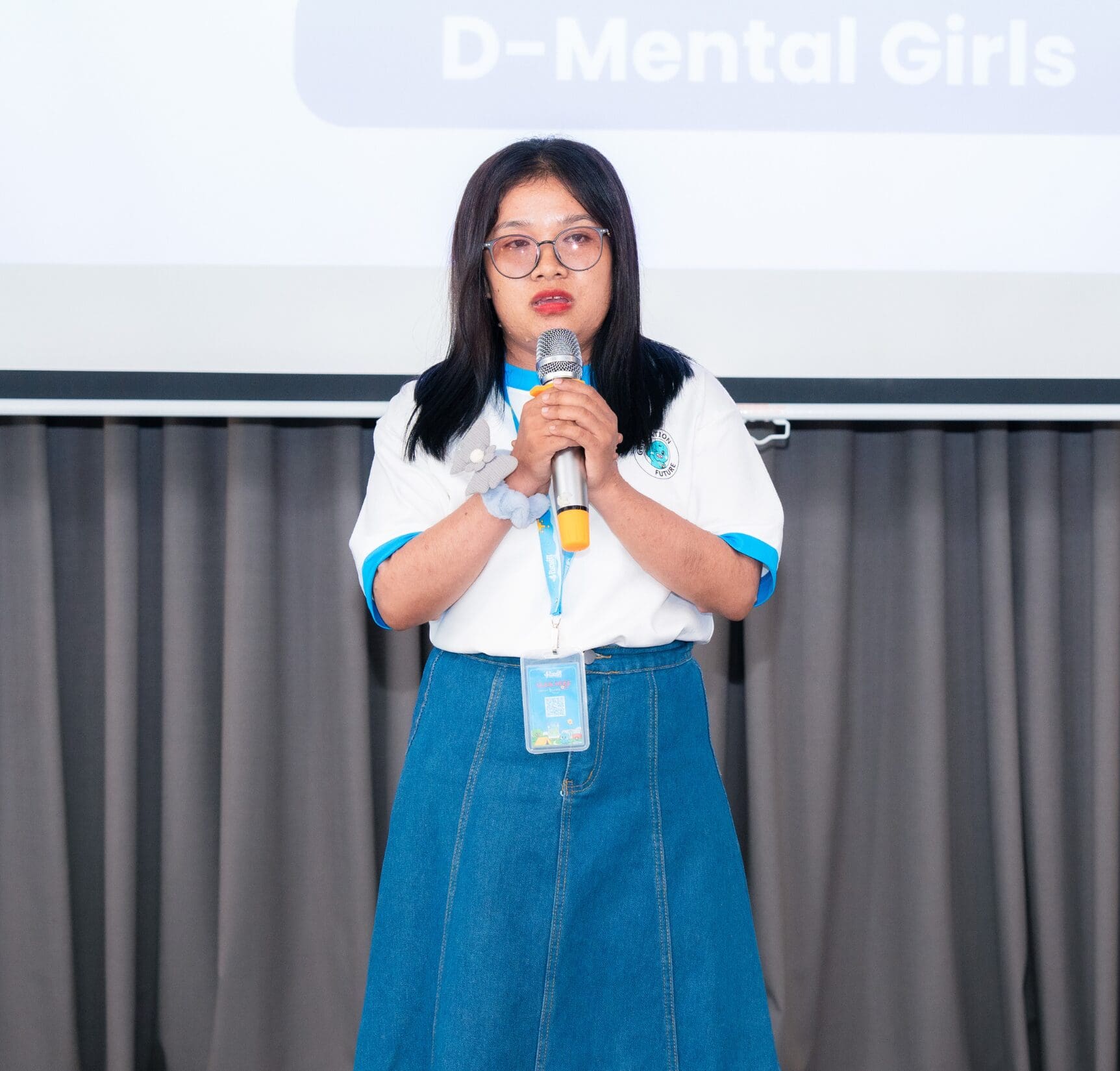 A person stands holding a microphone, wearing glasses, a white and blue shirt, and a blue skirt. A screen behind them displays the text "D-Mental Girls."