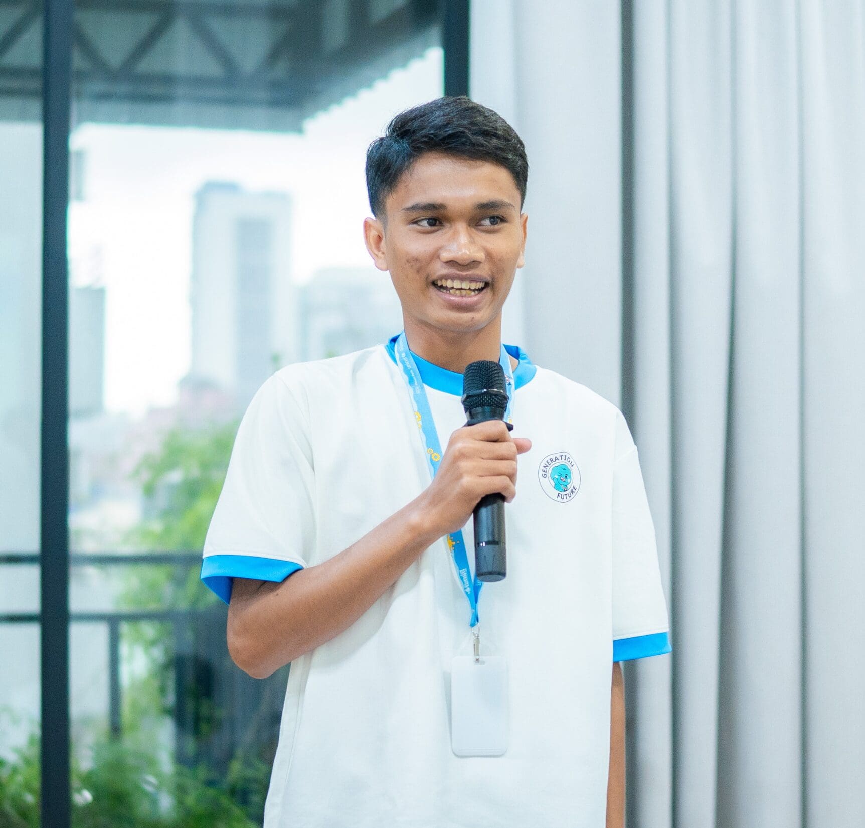 A person in a white and blue shirt speaks into a microphone indoors, smiling with a relaxed demeanor. A cityscape is visible through a window.