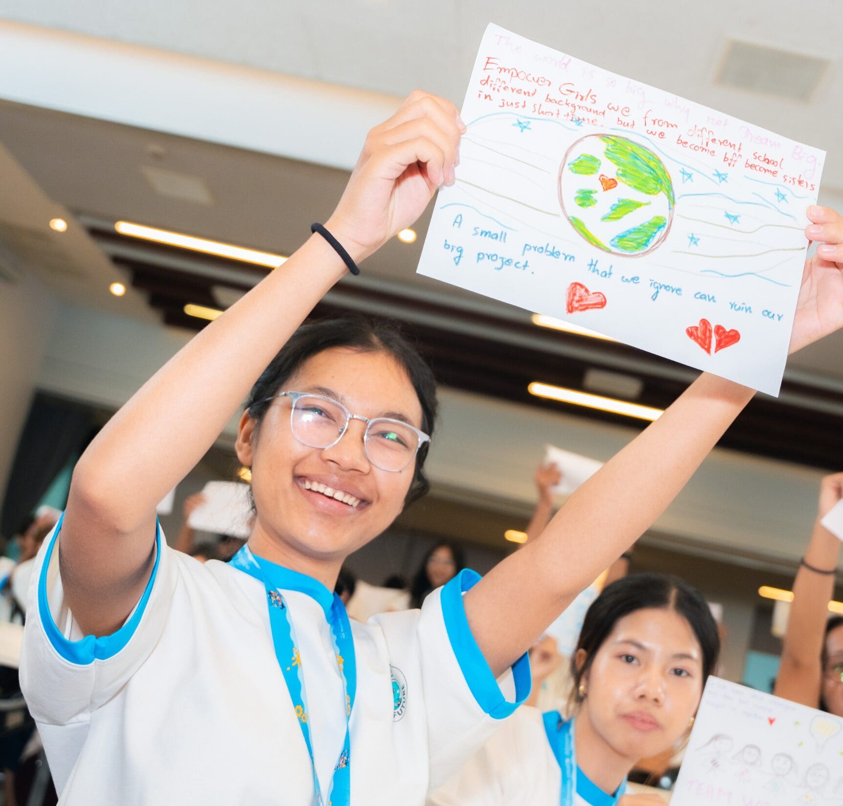 Smiling girl holds a colorful drawing of Earth and hearts, wearing glasses and a uniform, in a lively classroom, conveying empowerment and positivity.