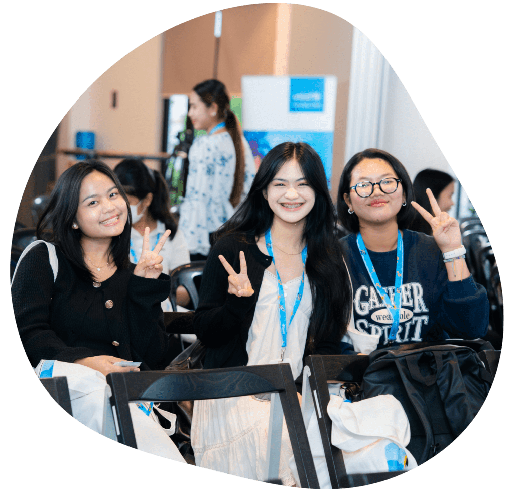 Three people sit smiling and making peace signs at an event. They wear lanyards and casual attire, creating a cheerful and relaxed atmosphere.