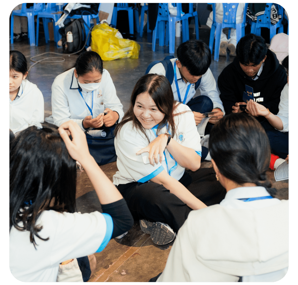 A group of students in white uniforms sit in a circle on the floor, smiling and engaging in an activity. The atmosphere is lively and cheerful.