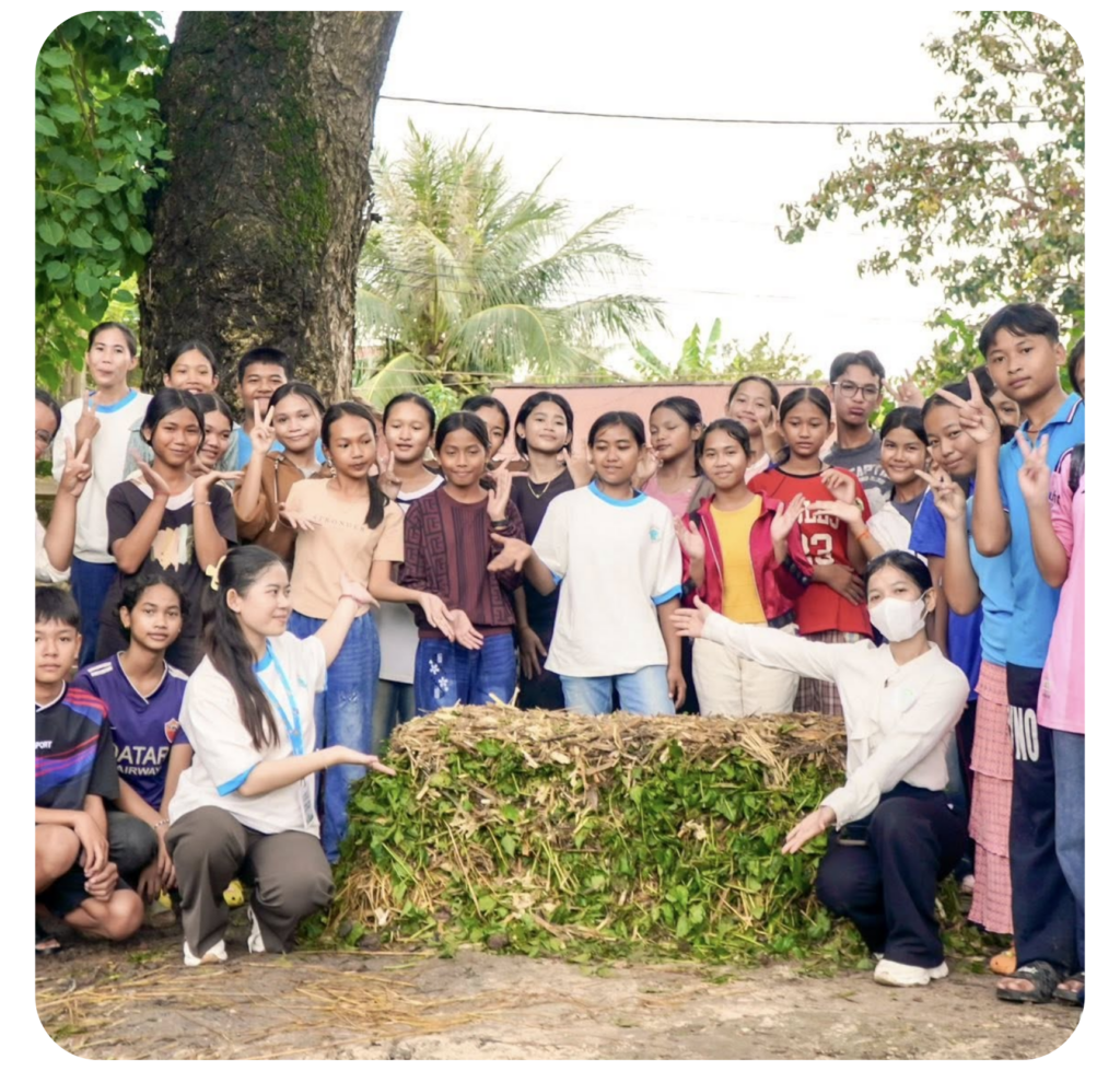 A group of smiling children and two adults gather outside, posing proudly around a rectangular compost pile made of leaves. The mood is joyful and communal.