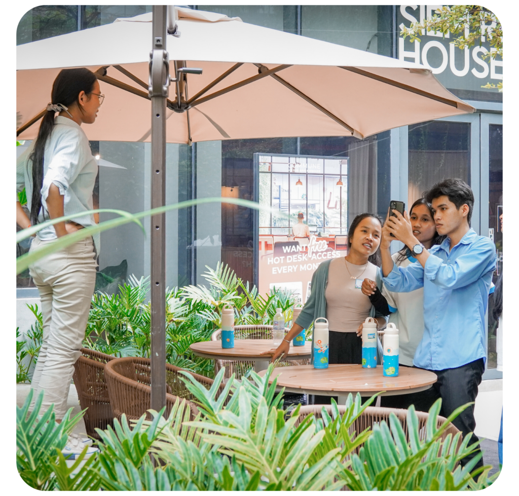 A group of four people at an outdoor café, engaged and smiling under a beige umbrella. They gather around round tables with plants and tumblers.