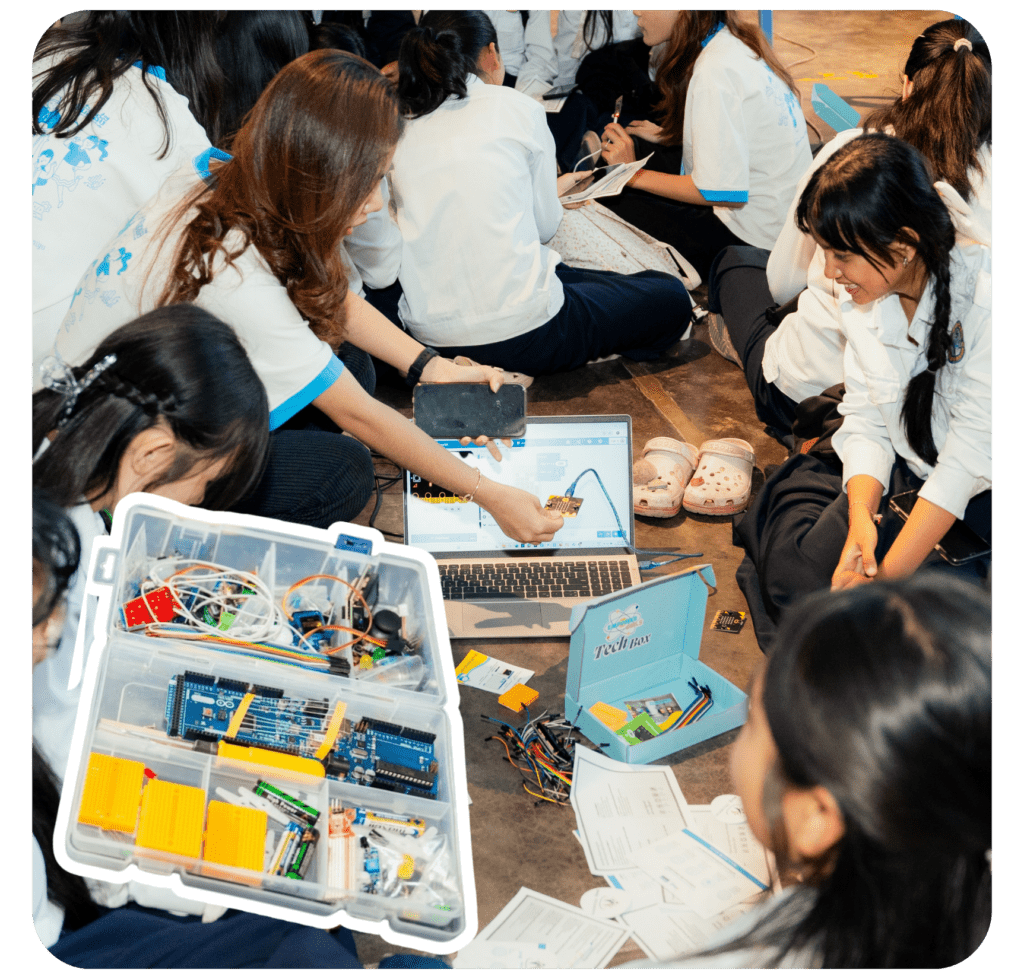 A group of students in uniforms sit on the floor, engaged in a hands-on electronics project. They use a laptop and components from an open toolkit.