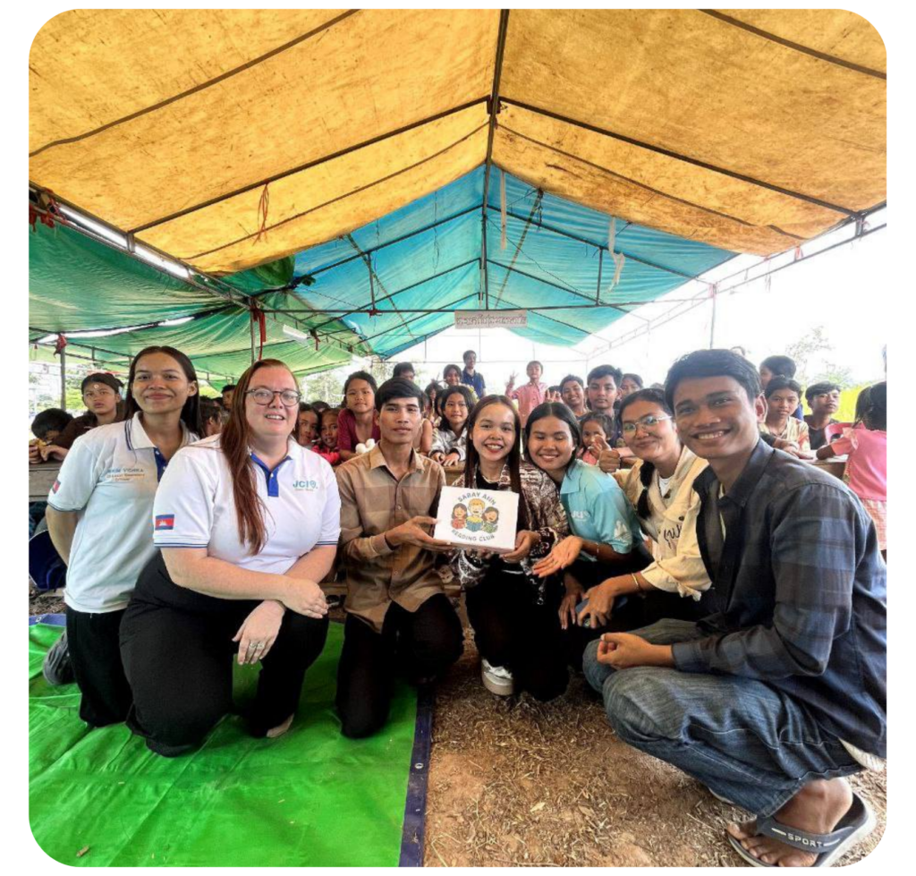 A group of smiling people pose under a large tent. They hold a drawing, surrounded by many others in the background. The mood is joyful and communal.