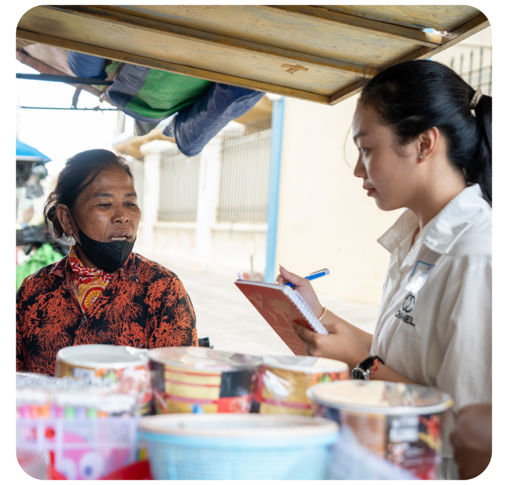 A woman in a patterned shirt, wearing a mask, talks to another woman holding a notebook and pen. They stand by a cart filled with goods, engaged in conversation.