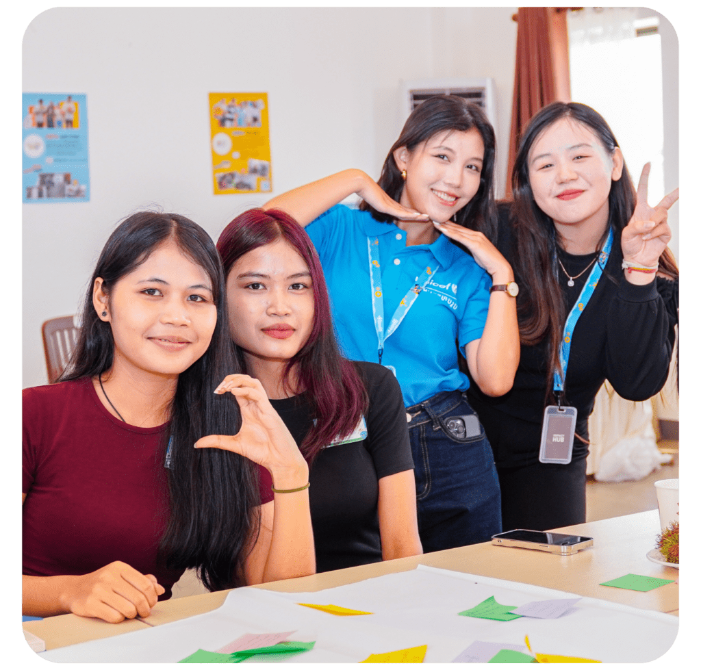 Four young women gather around a table, smiling and gesturing playfully. The setting is casual, with colorful posters in the background. The mood is cheerful and friendly.