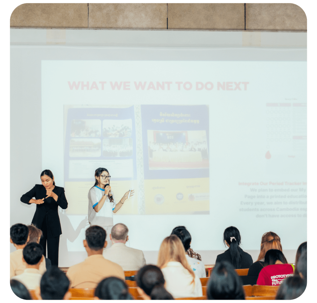 A woman speaks into a microphone, presenting to an audience while another signs in sign language. The slide reads "What We Want to Do Next."