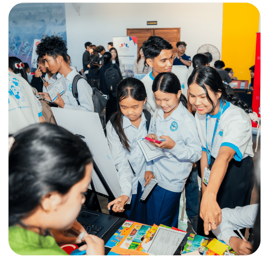 A group of young people in uniforms gather around a display booth at a busy event. They look engaged and curious, with one pointing and others taking notes. The atmosphere is lively and educational.