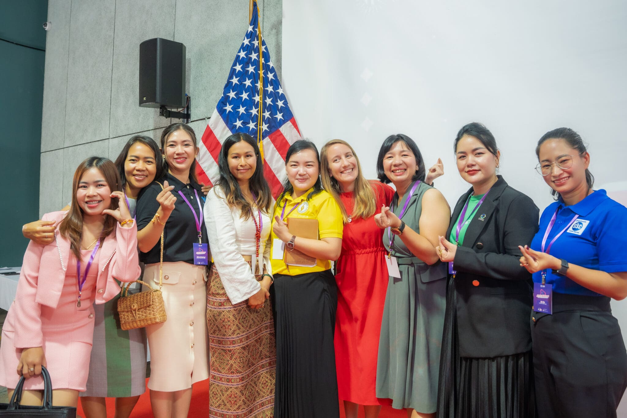 A group of smiling women stands together, making heart gestures with their fingers. An American flag is in the background, creating a joyful atmosphere.