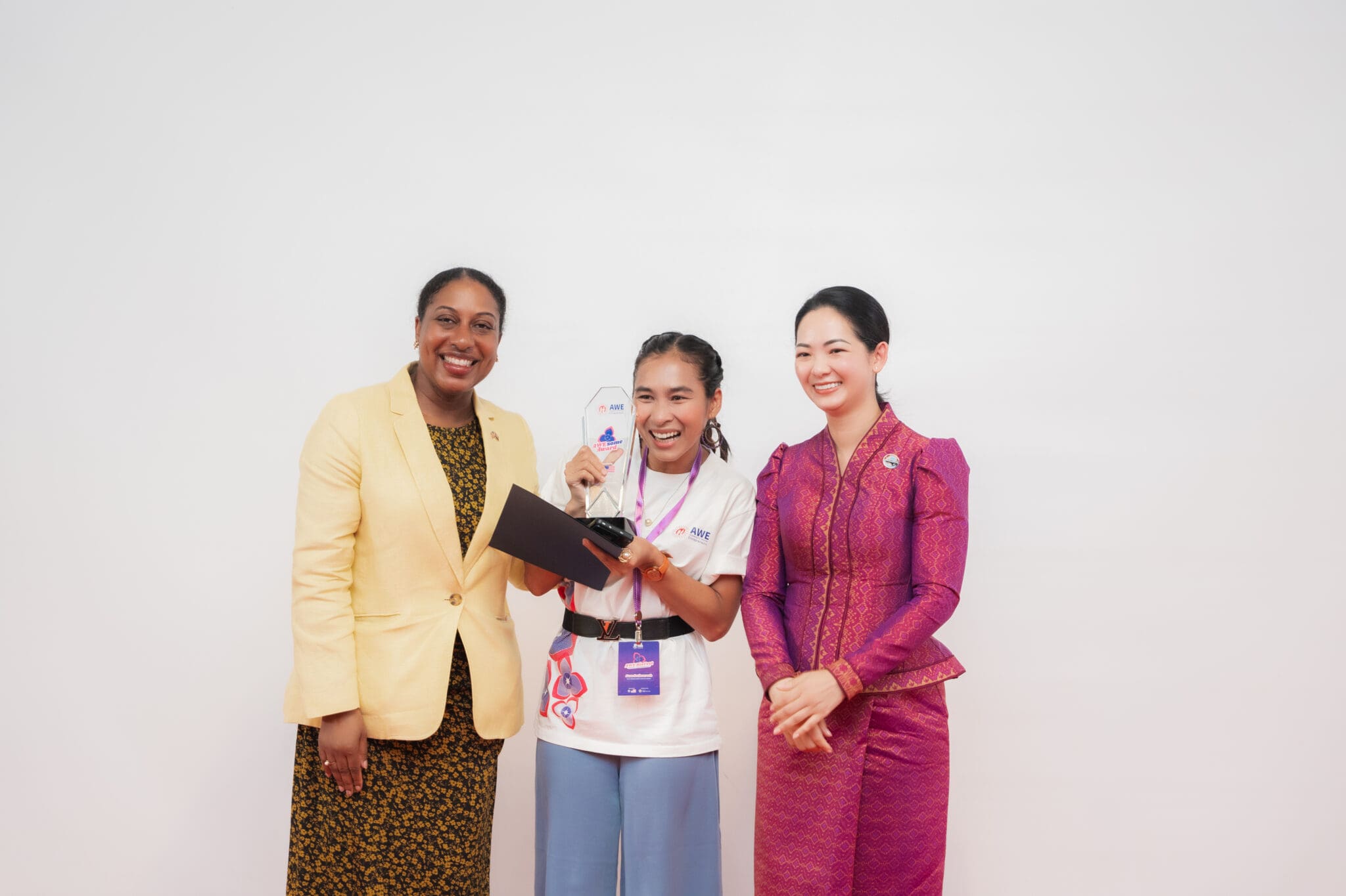 Three people smiling against a plain background; one holds an award. The tone is celebratory, with diverse attire indicating a formal event.