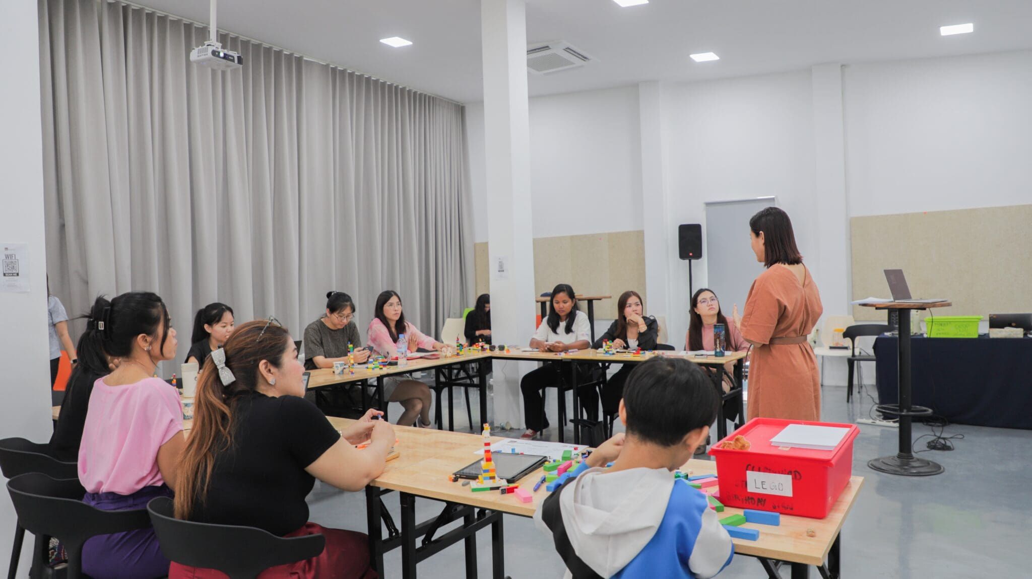 A group of people sit at tables in a bright classroom, attentively watching a woman standing and speaking. Art supplies are scattered on the tables, suggesting a creative activity.