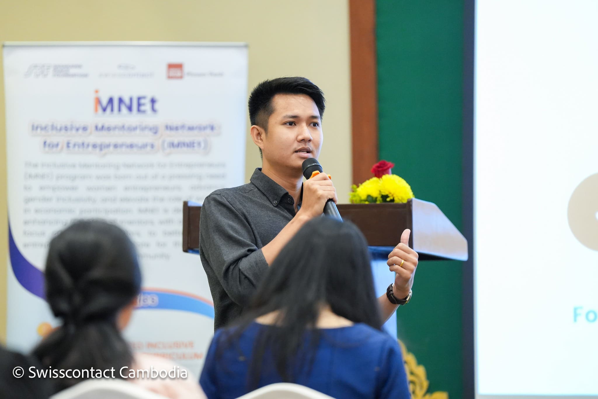 A man speaks into a microphone at a conference, standing near a podium with flowers. In the background, a blurred banner reads "Inclusive Mentoring Network." The atmosphere is professional and engaged.