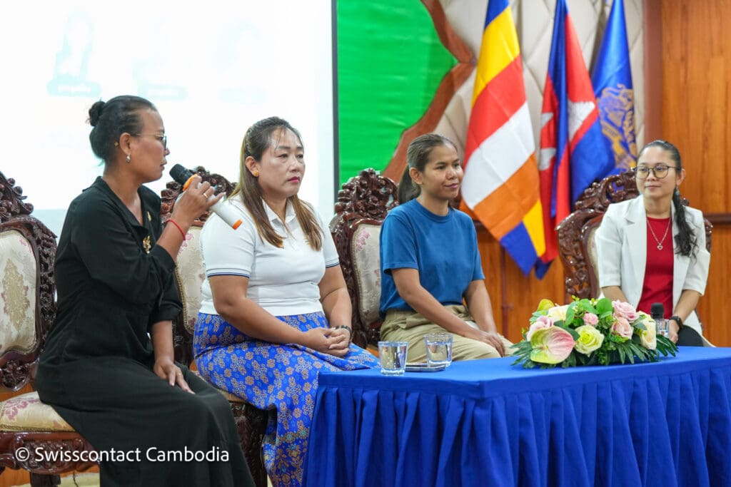 Four women sit on ornate chairs during a panel discussion. The woman on the left speaks into a microphone. Flags and a blue table with flowers are visible.