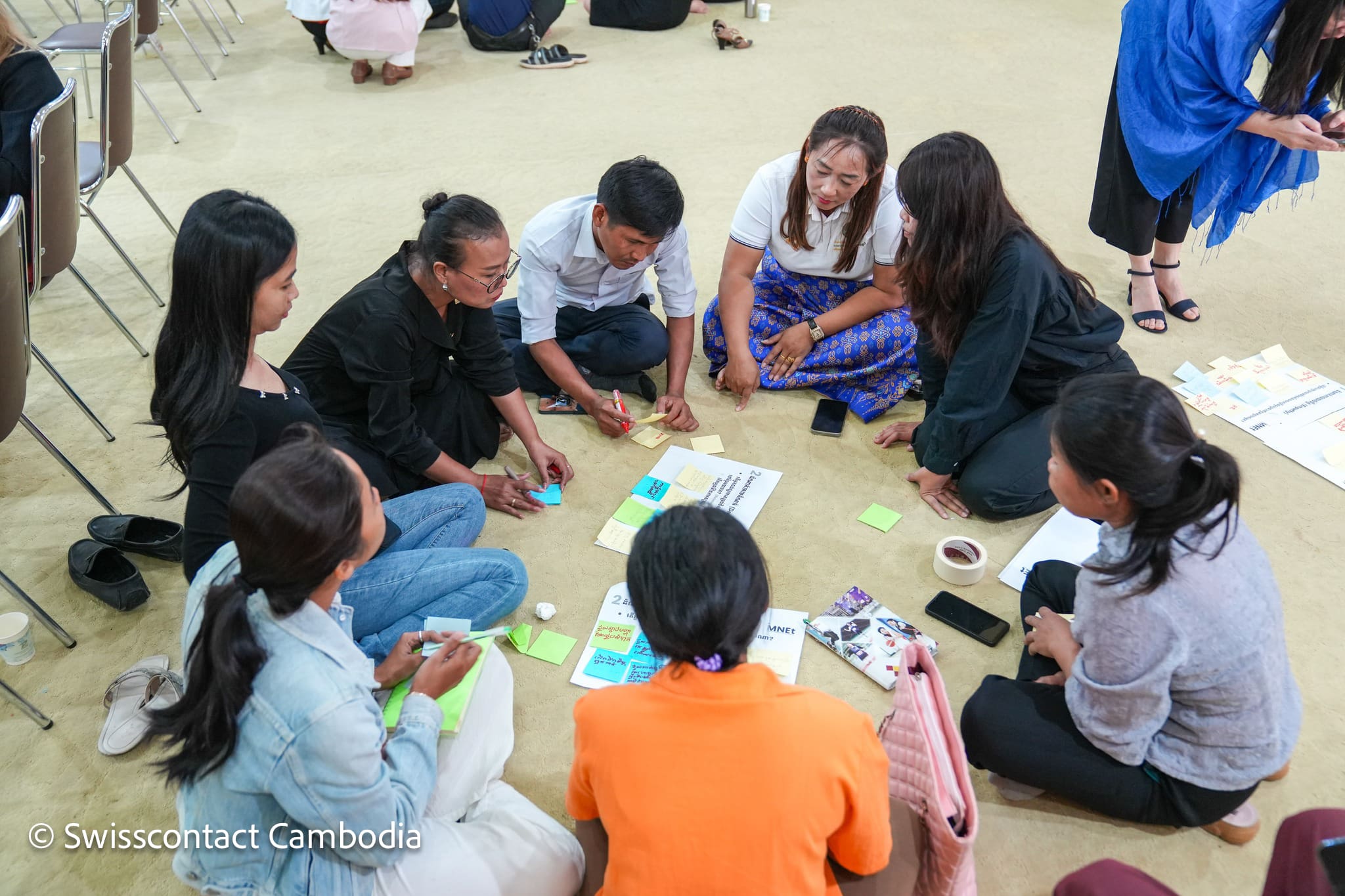 A group of people sit on the floor in a circle, actively discussing and writing on colorful sticky notes and paper. The setting is collaborative and focused.