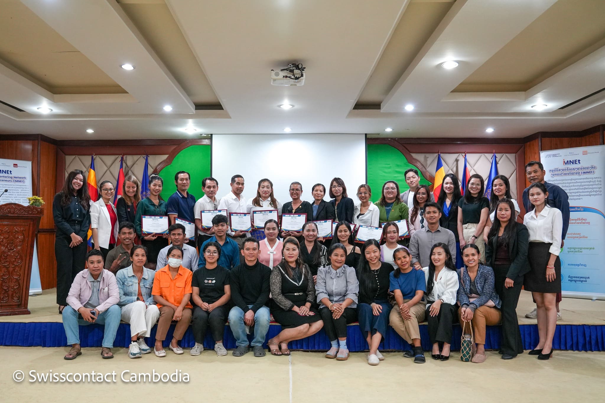 A group of 36 people poses on a stage in an auditorium, smiling proudly. Some hold certificates. Background features colorful flags and banners.