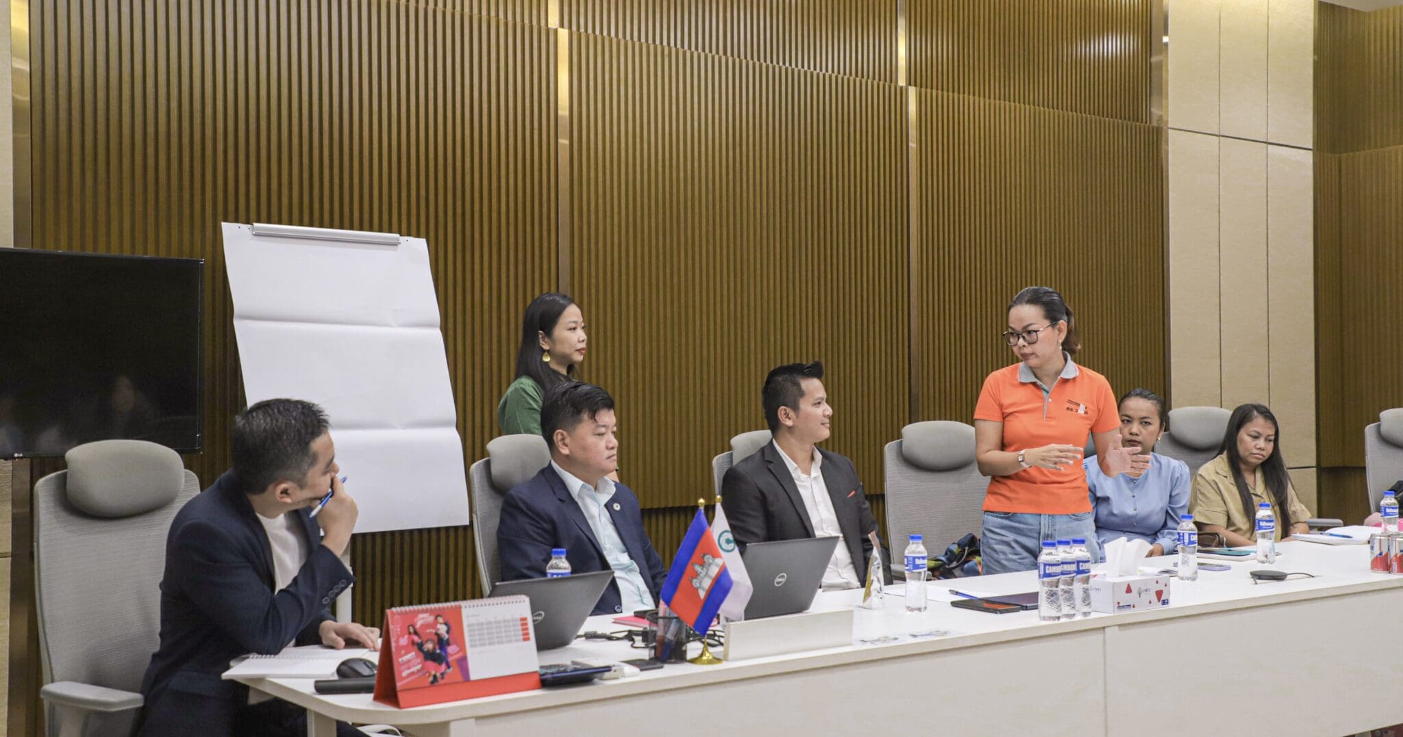 A group of six people in a conference room, engaged in a discussion. One person stands, speaking to the others who are seated around a long table with laptops and papers.