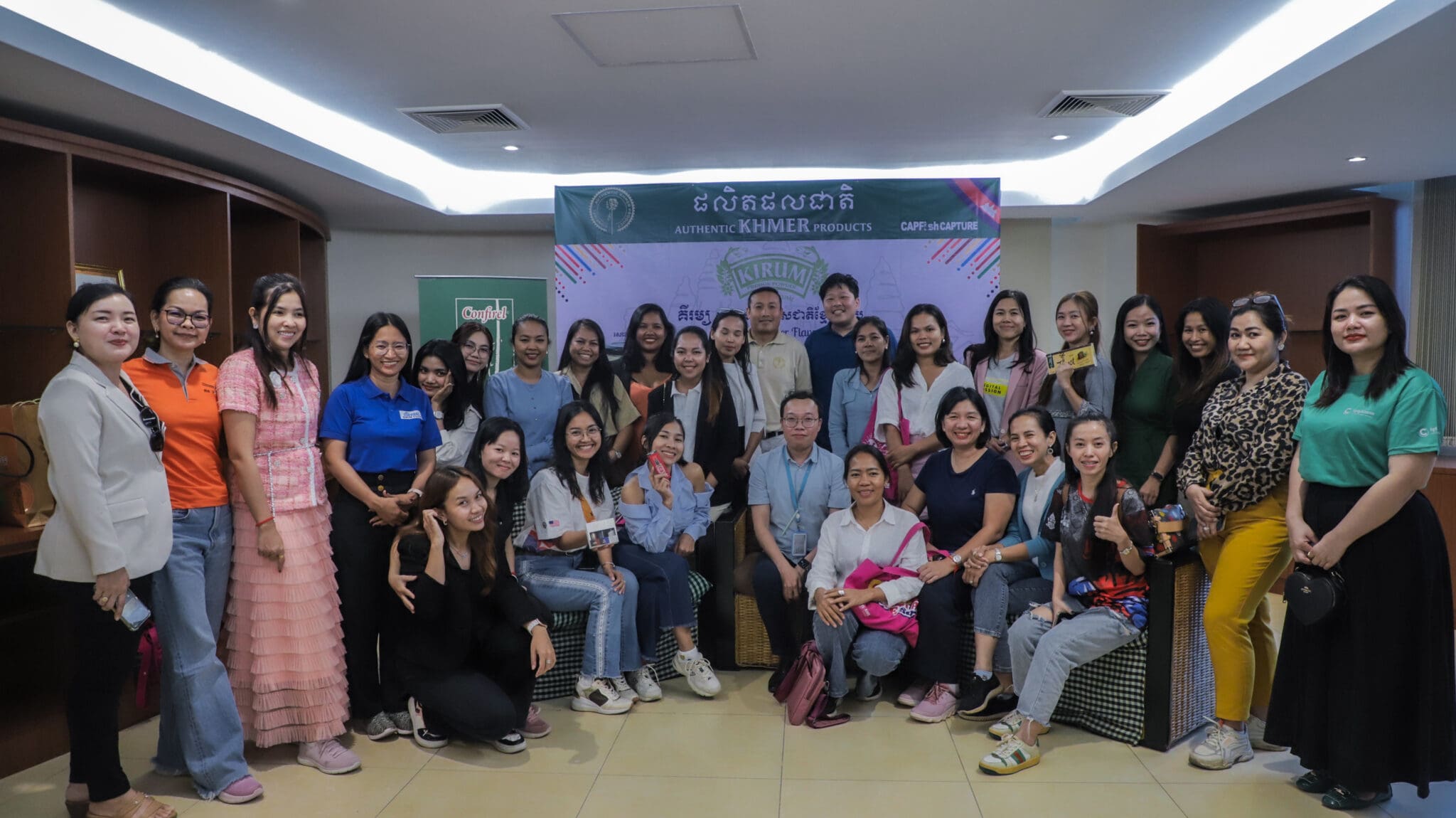 A group of 26 people, mostly women, poses indoors in front of a banner labeled "Authentic Khmer Products." They appear cheerful and relaxed.