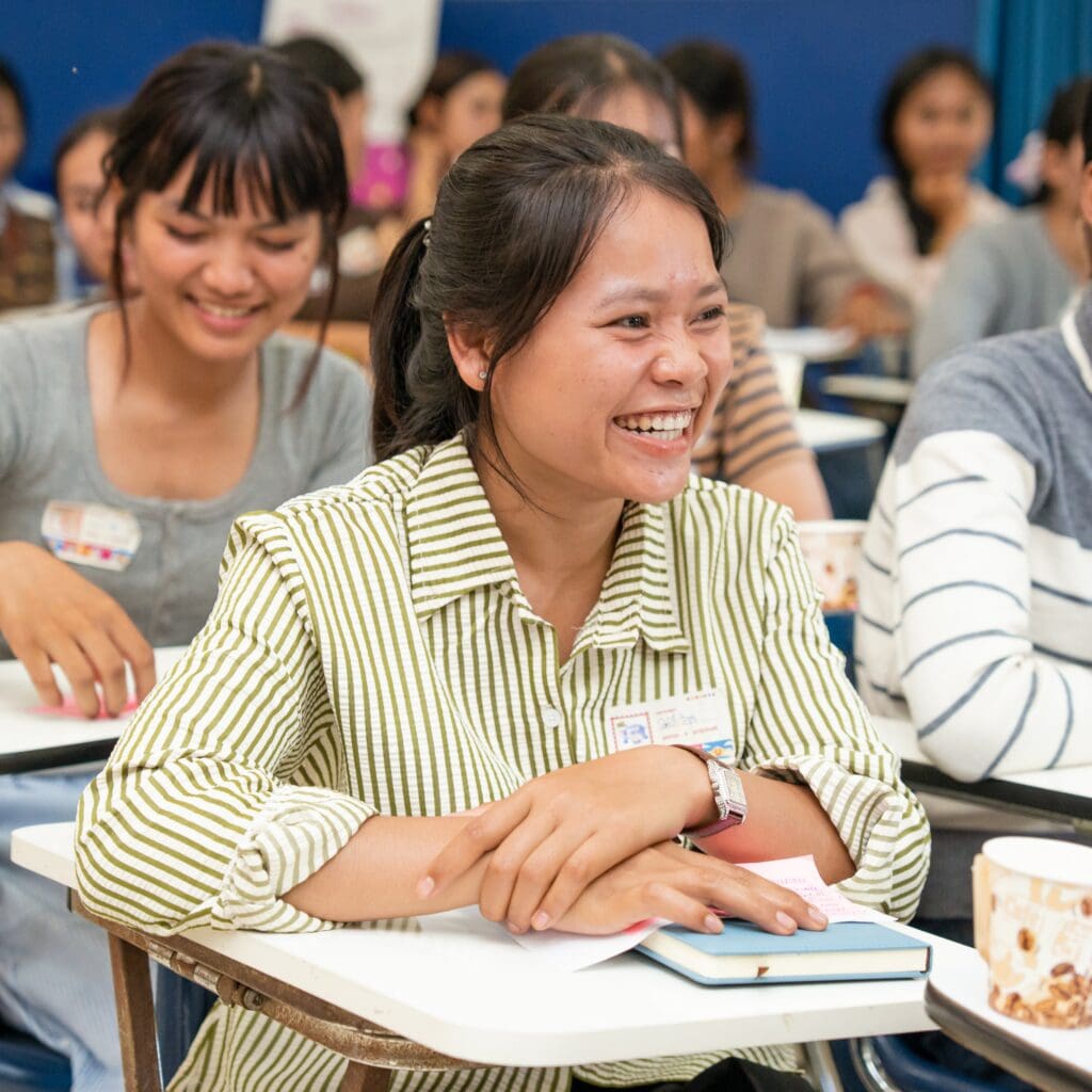 A woman in a striped shirt sits at a desk, smiling and looking engaged in a classroom setting. Other students are seen in the background, also smiling.