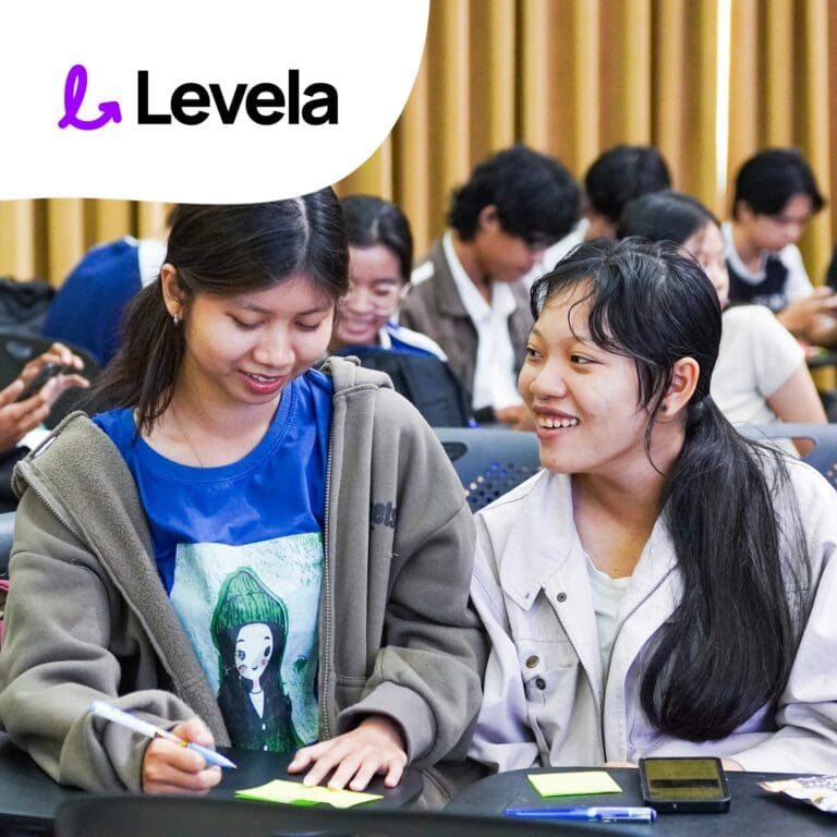 Two young women sit at a table, writing on sticky notes and smiling, with other people working in the background. The Levela logo is displayed in the top left corner.