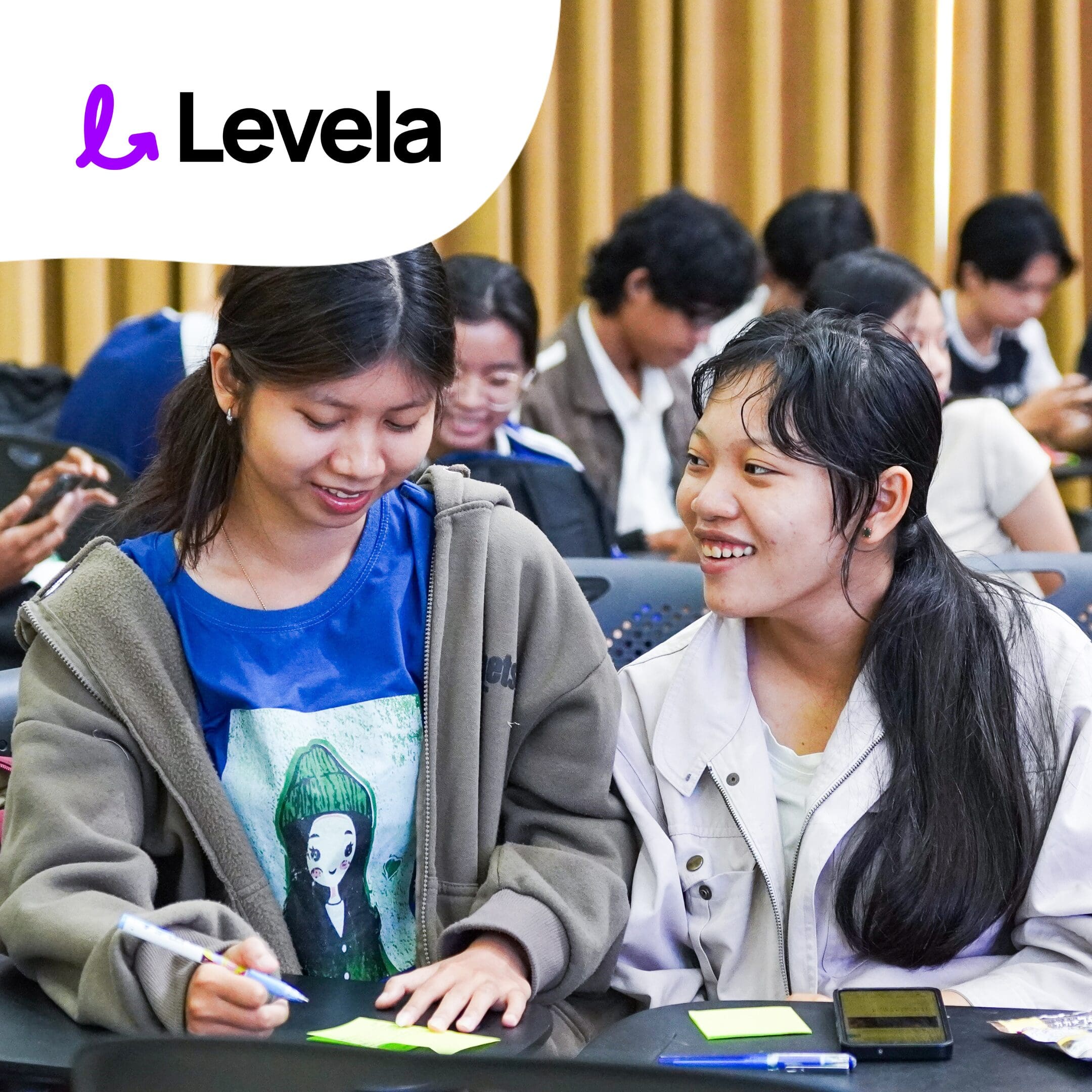 Two young women sit at a table, writing on sticky notes and smiling, with other people working in the background. The Levela logo is displayed in the top left corner.