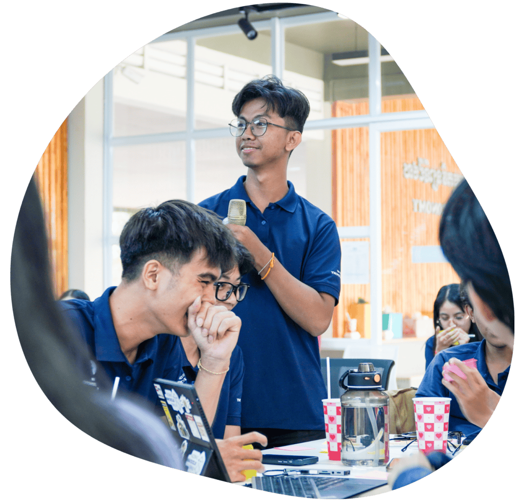 A group of students in a classroom setting, with one student standing and holding a drink, while others are engaged in conversation and working on laptops.