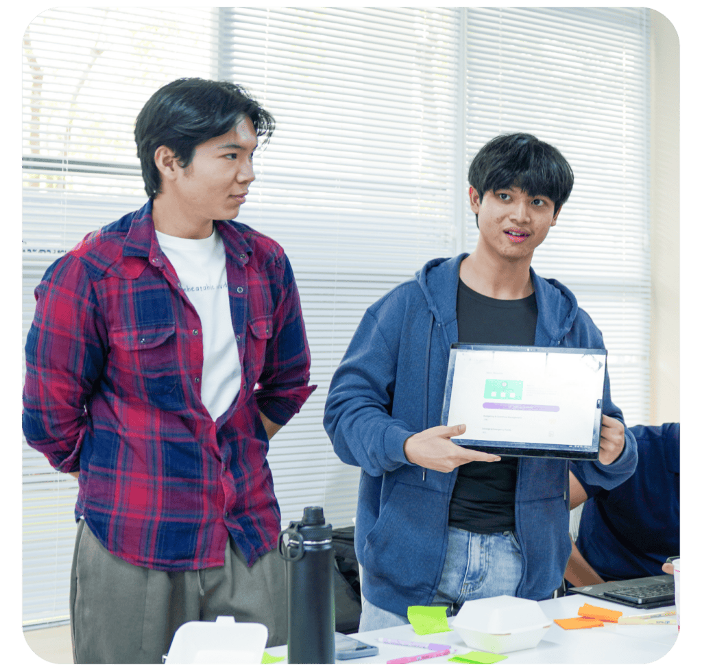 Two young men are indoors. One is wearing a plaid shirt and looks on as the other holds up a tablet displaying text, possibly presenting to a group.